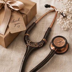 Puede incluir: Un estetoscopio negro y oro rosa con una etiqueta de nombre de alambre personalizada que dice Justin con un pequeño símbolo de corazón. El instrumento médico se muestra junto a una caja de regalo marrón con una cinta beige y una etiqueta thank you.