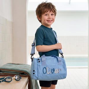 Könnte beinhalten: Eine hellblaue Sporttasche mit dem Namen Oliver, der in einer dunkelblauen Schreibschrift auf der Vorderseite eingestickt ist. Die Tasche verfügt über zwei Tragegriffe, einen abnehmbaren Schultergurt mit schwarzen Beschlägen und einen seitlichen Reißverschluss.