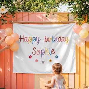 May include: A white fabric banner featuring the colourful text Happy birthday Sophie and small flower accents. The banner is decorated with clusters of pastel balloons on both sides and is displayed against a vertical wooden slat wall.
