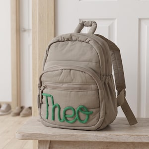 May include: A taupe quilted backpack featuring a top handle, adjustable shoulder straps, and a front zippered pocket. The name Theo is embroidered on the front pocket in a bright green, thick, rope-like script font with a textured appearance.