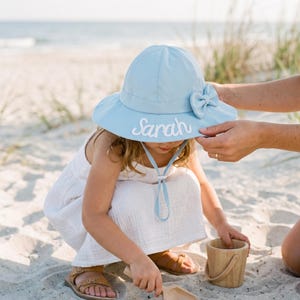 May include: A light blue sun hat featuring a wide brim, a decorative side bow, and a chin strap with an adjustable toggle. The name Sarah is embroidered in white script on the front of the brim. The hat is constructed from a solid fabric.