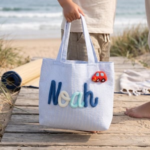 May include: A light blue and white seersucker tote bag featuring the name Noah in raised, dark blue and light blue yarn lettering, with a small, red, felt-like car appliqué attached to the upper right corner of the bag's front panel.
