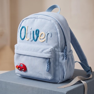 May include: A light blue corduroy backpack featuring the name Oliver embroidered in blue and cream rope-style lettering across the top. A small red and blue car applique is attached to the front pocket, which has matching blue braided zipper pulls.