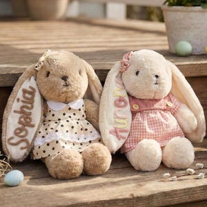 Peut inclure: Deux lapins en peluche, un beige et un crème, sont assis sur des marches en bois. Le lapin beige porte une robe à pois avec le nom "Sophia" sur son oreille. Le lapin crème a une robe à carreaux roses et le nom "Aurora" sur son oreille. Des œufs de Pâques et un panier de fleurs sont à proximité.