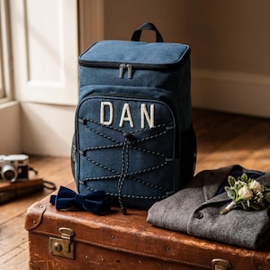 May include: A blue denim backpack featuring the name DAN in white letters on the front pocket, accented with black bungee cord lacing. The backpack has a top handle and dual zippers, providing a structured storage solution for personal items.