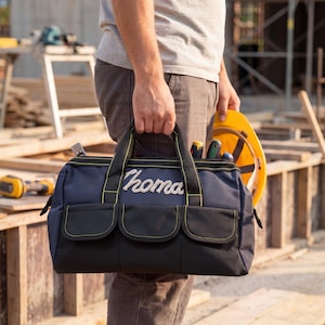 May include: A navy and black tool bag with the name "Thoma" embroidered in white. The bag features three front pockets and yellow stitching. A yellow hard hat and tools are visible.