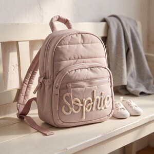 Puede incluir: Una mochila acolchada de color rosa polvoriento con asa superior, correas ajustables para los hombros y un bolsillo frontal con cremallera. El nombre Sophie está bordado en grandes letras cursivas de color crema tipo cuerda en el bolsillo.