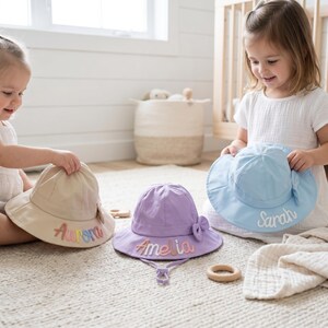 May include: Three sun hats in beige, lavender, and light blue. The beige hat features the name Aurora in colorful embroidery, the lavender hat displays the name Amelia, and the light blue hat shows the name Sarah in white cursive embroidery.