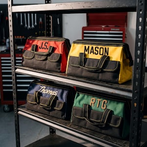 May include: Four personalised tool bags in red, yellow, blue, and green, each with a different name embroidered on the top. The bags have black bases, multiple pockets, and black handles. They are displayed on a metal shelf.