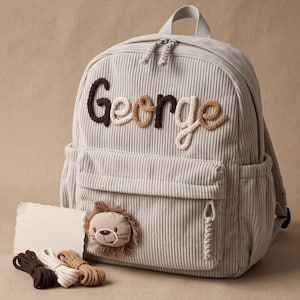 Puede incluir: Una mochila de pana gris claro con el nombre George bordado en letras de cuerda gruesas y texturizadas en tonos marrón, crema y bronceado. Un pequeño parche de cara de león de felpa está unido al bolsillo delantero con tirador trenzado.