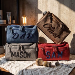 May include: Four rectangular toiletry bags in blue, brown, gray, and red, each featuring a zippered closure and a name embroidered in thick yarn: DAD, Oliver, MASON, and SAM. A wooden shaving brush and a metal safety razor sit in the foreground.