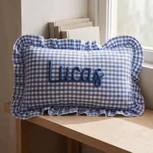 Pu&ograve; includere: Un cuscino rettangolare con motivo a quadretti vichy blu e bianchi e un bordo arricciato. Il nome Lucas &egrave; ricamato in corsivo blu scuro al centro del cuscino, utilizzando una texture spessa e in rilievo simile a una corda.