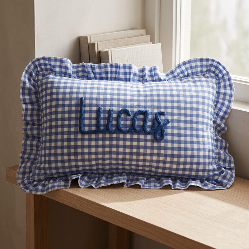 Pu&ograve; includere: Un cuscino rettangolare con motivo a quadretti vichy blu e bianchi e un bordo arricciato. Il nome Lucas &egrave; ricamato in corsivo blu scuro al centro del cuscino, utilizzando una texture spessa e in rilievo simile a una corda.