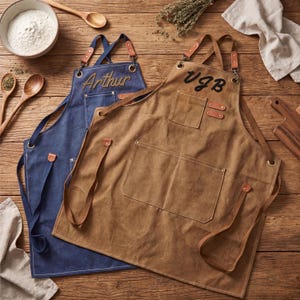May include: Two aprons on a wooden surface. One is navy blue with "Arthur" embroidered in gold, the other is brown with "VGB" embroidered in black. Both have pockets, leather accents, and adjustable straps.