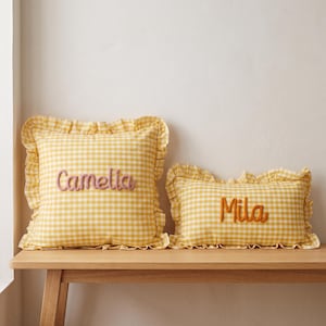 May include: Two square and rectangular throw cushions featuring a yellow and white gingham check pattern with ruffled edges. The square cushion has the name Camelia embroidered in pink, and the rectangular cushion has the name Mila in orange.