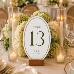May include: An oval table number sign labeled 13 with the initials Z & R and the date 10.20.202x, featuring a sage green border on a white background. The sign is held by a rectangular wooden base and is surrounded by white hydrangea flowers.
