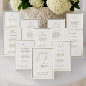 May include: A collection of white rectangular wedding seating chart cards with sage green borders and elegant script text. Each card displays a table number from one to nine, a list of names, and the text Please Find Your Seat, Sofia & Liam, August 22.20XX.