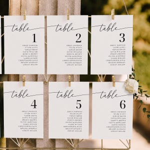 Puede incluir: Seis tarjetas rectangulares blancas de distribución de mesas de boda con texto en negro, cada una etiquetada como table 1 a 6 en fuente cursiva seguida de una lista de nombres. Las tarjetas están sujetas a una rejilla dorada con clips.