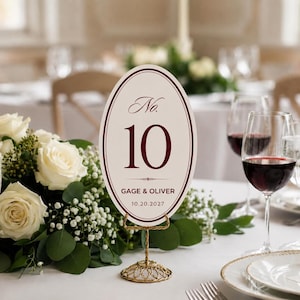 Puede incluir: Una tarjeta de número de mesa ovalada con fondo crema y borde rojo oscuro. La tarjeta muestra el texto No. 10, los nombres GAGE & OLIVER y la fecha 10.20.2027. Se mantiene en posición vertical mediante un soporte decorativo de alambre dorado.