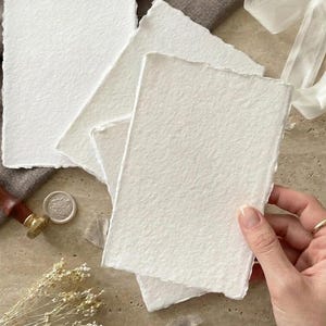 Puede incluir: Varias hojas de papel blanco texturizado, con bordes irregulares, dispuestas sobre una superficie neutra. El papel tiene un tacto ligeramente rugoso y org&aacute;nico. Una mano sostiene una pila de papel, lo que sugiere su uso para papeler&iacute;a o proyectos art&iacute;sticos.