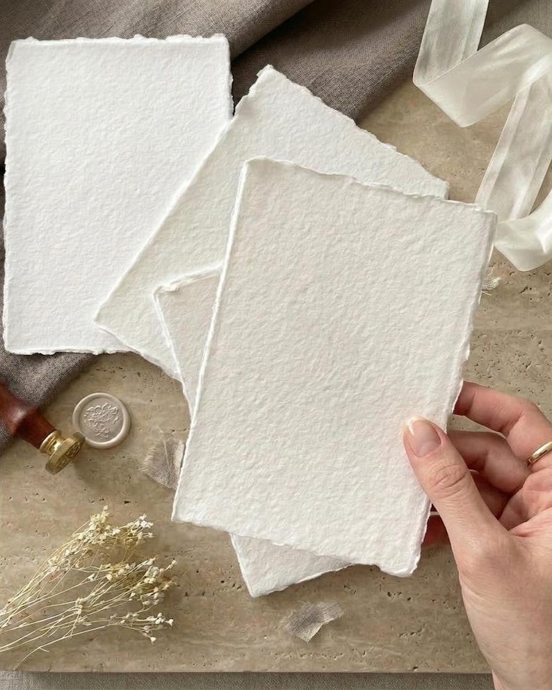 Puede incluir: Varias hojas de papel blanco texturizado, con bordes irregulares, dispuestas sobre una superficie neutra. El papel tiene un tacto ligeramente rugoso y org&aacute;nico. Una mano sostiene una pila de papel, lo que sugiere su uso para papeler&iacute;a o proyectos art&iacute;sticos.