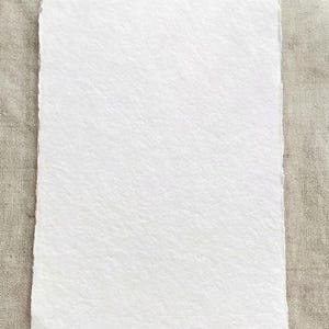 Puede incluir: Una hoja rectangular de papel blanco hecho a mano con una superficie texturizada y bordes irregulares. El papel se muestra sobre un fondo de tonos neutros, resaltando su color y textura. Ideal para proyectos art&iacute;sticos o caligraf&iacute;a.