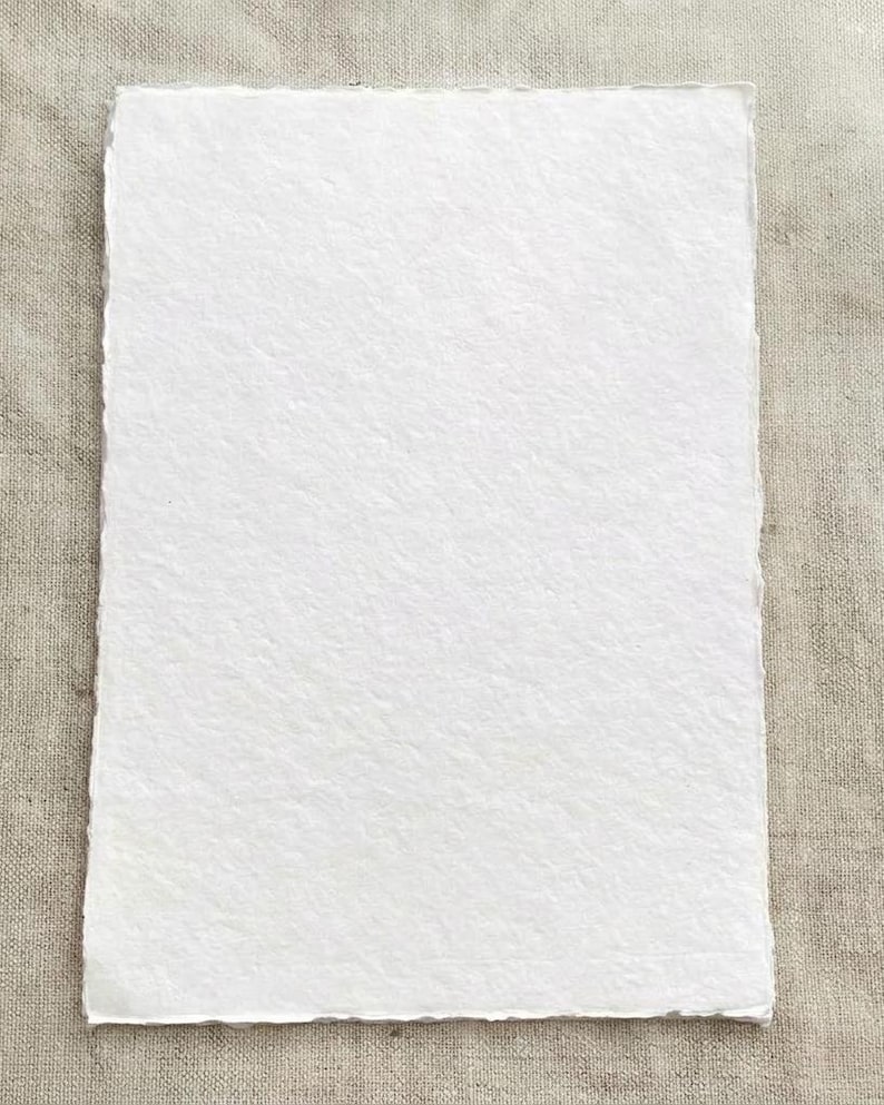 Puede incluir: Una hoja rectangular de papel blanco hecho a mano con una superficie texturizada y bordes irregulares. El papel se muestra sobre un fondo de tonos neutros, resaltando su color y textura. Ideal para proyectos art&iacute;sticos o caligraf&iacute;a.