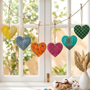 Peut inclure: Six décorations en forme de cœur en tissu suspendues à une ficelle de jute. Les cœurs présentent différents motifs et couleurs : jaune, bleu, orange, rose, turquoise et vert. Ils sont suspendus devant une fenêtre.