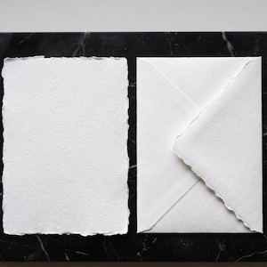 Könnte beinhalten: Ein weißes Briefpapier-Set mit einer strukturierten Papierkarte mit zerrissenen Kanten und einem passenden Umschlag. Das Briefpapier wird auf einer dunklen, marmorierten Oberfläche präsentiert, wodurch eine minimalistische Ästhetik entsteht. Ideal für Einladungen oder persönliche Notizen.