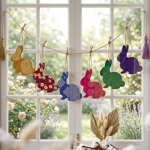 May include: A colourful Easter bunny garland with six fabric bunnies suspended in front of a window. The bunnies are in gold, red, blue, pink, green, and purple, each with unique patterns. Tassels decorate the ends of the twine.