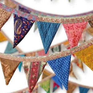 May include: A colorful fabric bunting with triangular flags. The flags feature various patterns and colors, including paisley, floral, and geometric designs. The bunting is strung together with a decorative trim, creating a festive and bohemian aesthetic.