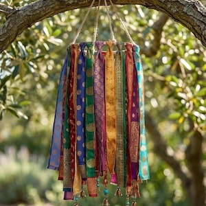 May include: A hanging mobile featuring multiple vertical strips of colorful patterned fabric in shades of blue, green, red, gold, and purple. The fabric strips are attached to a circular frame with twine and accented with small metal bells and beads.