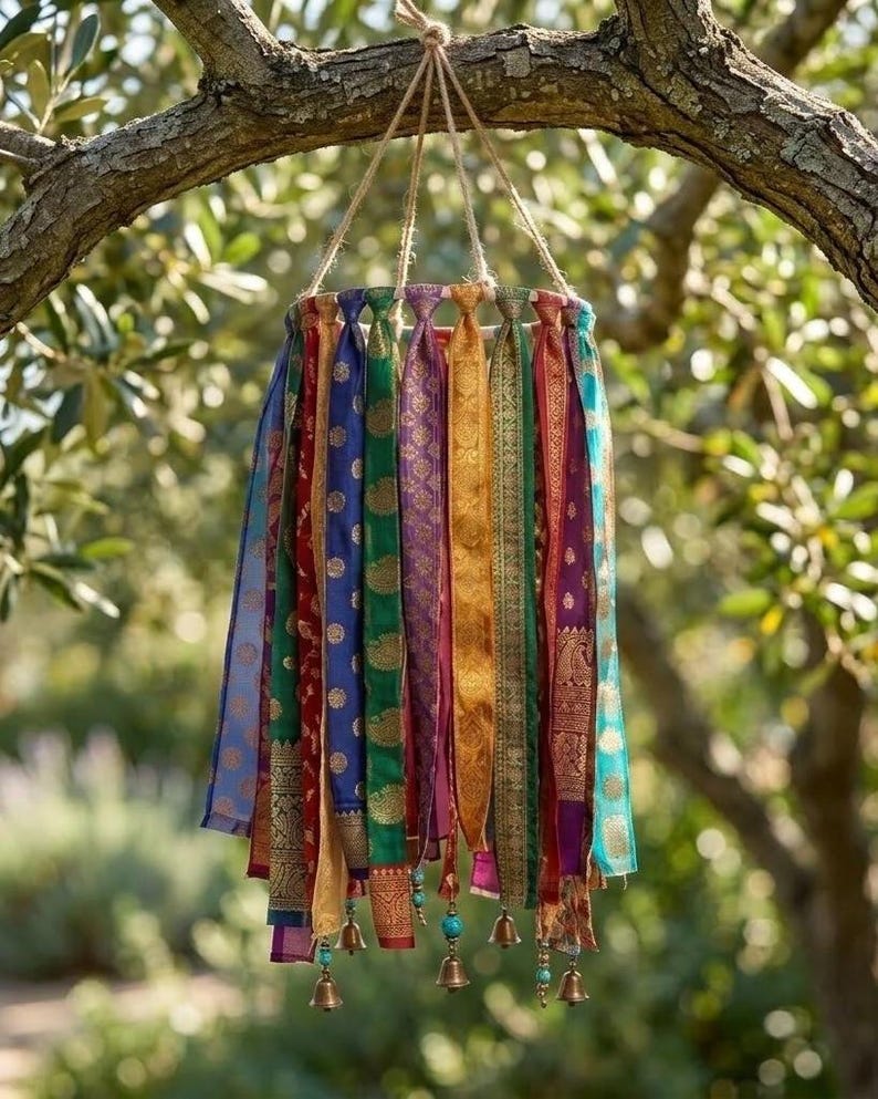 May include: A hanging mobile featuring multiple vertical strips of colorful patterned fabric in shades of blue, green, red, gold, and purple. The fabric strips are attached to a circular frame with twine and accented with small metal bells and beads.