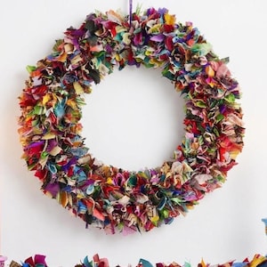 May include: A colorful, handmade wreath made of fabric scraps in various colors, including red, purple, green, and yellow. The wreath is circular and hangs from a purple string. The wreath is a decorative item.