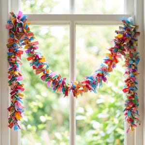 May include: A colorful fabric garland hangs in front of a window. The garland is made of small, rectangular fabric pieces in various colors, including pink, blue, yellow, and orange. The garland is a decorative item.