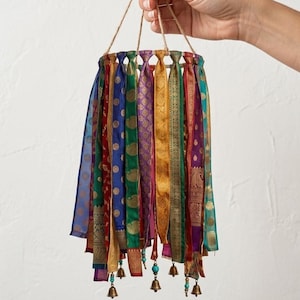 May include: A circular hanging mobile featuring numerous vertical fabric ribbons in vibrant shades of blue, green, red, gold, and purple, each adorned with intricate patterns. Small metallic bells and turquoise beads hang from the bottom edges.