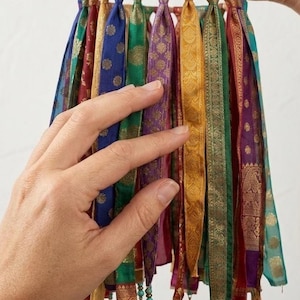 May include: A hanging mobile featuring vertical strips of colorful patterned fabric in shades of blue, green, purple, and gold. The fabric strips are attached to a circular frame and decorated with small metal bells and turquoise beads.