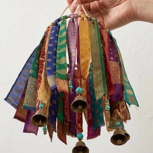 May include: A circular mobile featuring multiple hanging strips of colorful patterned fabric in shades of blue, green, purple, red, and gold. Several brass bells are suspended from the fabric strips, accented with small turquoise beads.