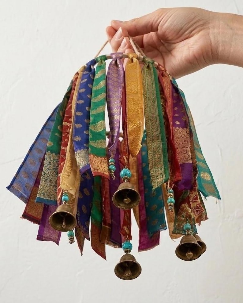 May include: A circular mobile featuring multiple hanging strips of colorful patterned fabric in shades of blue, green, purple, red, and gold. Several brass bells are suspended from the fabric strips, accented with small turquoise beads.