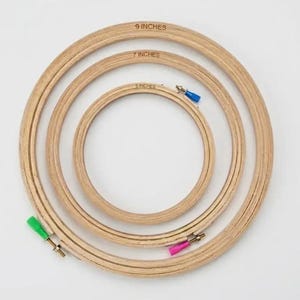May include: Three circular wooden embroidery hoops nested inside one another. The hoops are labeled 9 inches, 7 inches, and 5 inches. Each hoop features a metal tightening screw with a colorful plastic cap in green, pink, or blue.