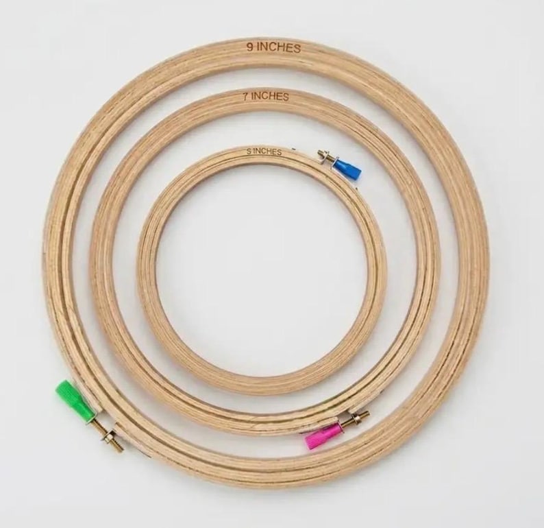 May include: Three circular wooden embroidery hoops nested inside one another. The hoops are labeled 9 inches, 7 inches, and 5 inches. Each hoop features a metal tightening screw with a colorful plastic cap in green, pink, or blue.