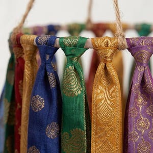 May include: A circular mobile featuring multiple hanging fabric strips in vibrant colors including blue, green, gold, red, and purple. Each strip displays a metallic gold woven pattern and is knotted around a circular frame suspended by jute twine.