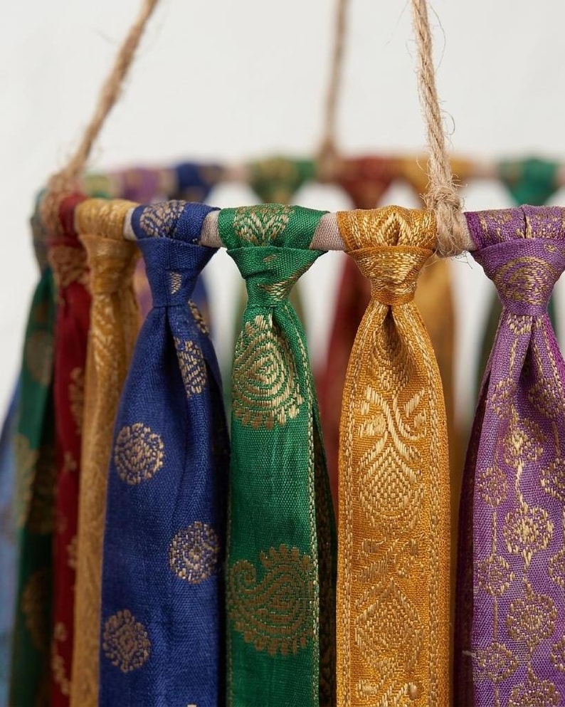 May include: A circular mobile featuring multiple hanging fabric strips in vibrant colors including blue, green, gold, red, and purple. Each strip displays a metallic gold woven pattern and is knotted around a circular frame suspended by jute twine.