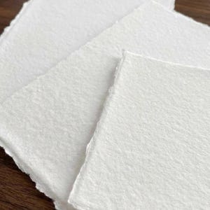 Puede incluir: Primer plano de varias hojas de papel blanco texturizado hechas a mano con bordes irregulares. El papel tiene una superficie ligeramente rugosa y est&aacute; dispuesto en un patr&oacute;n en capas, lo que sugiere un suministro de artesan&iacute;a o arte.
