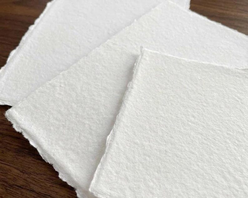 Puede incluir: Primer plano de varias hojas de papel blanco texturizado hechas a mano con bordes irregulares. El papel tiene una superficie ligeramente rugosa y est&aacute; dispuesto en un patr&oacute;n en capas, lo que sugiere un suministro de artesan&iacute;a o arte.