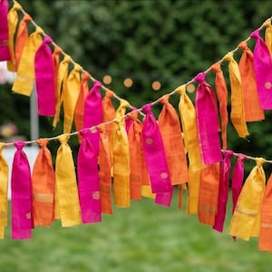 May include: A vibrant garland of fabric ribbons in shades of pink, orange, and yellow. The decorative string is suspended outdoors, perfect for celebrations, with a blurred green backdrop.