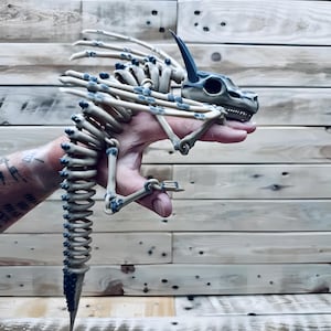 May include: A beige and blue dragon skeleton figurine. The dragon has a long, segmented tail and a detailed skull with a single horn. The skeleton is held in a hand against a wooden background.