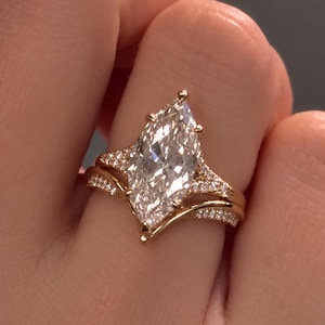 Peut inclure: Une bague en or avec une grande pierre centrale taille marquise fixée par des griffes. L'anneau principal et l'alliance incurvée assortie sont tous deux ornés de petites pierres serties pavé sur les côtés, créant un aspect superposé.
