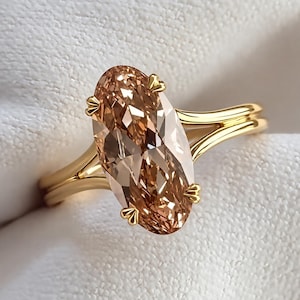 Peut inclure: Une bague en or ornée d'une grande pierre précieuse ovale de couleur champagne, maintenue par quatre griffes. L'anneau est divisé en deux fils d'or parallèles qui se rejoignent à la base du sertissage, offrant un aspect poli et métallique.