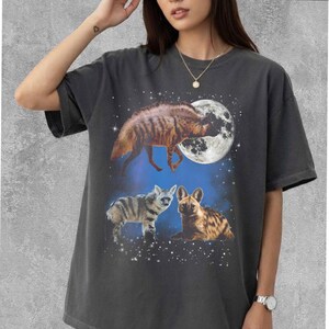 May include: Dark gray t-shirt featuring a graphic of hyenas under a full moon and starry sky. The design includes three hyenas in different poses, with the largest one in the center. The shirt is a relaxed fit.